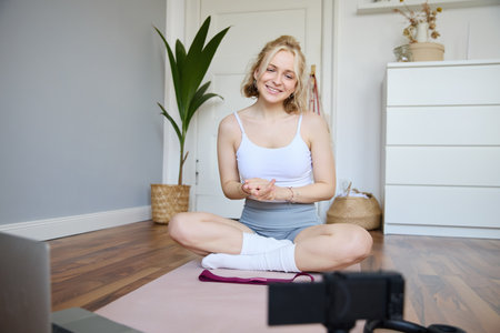 Young female athlete, fitness instructor woman sits on floor rubber mat, recording video on digital camera, showing how to workout, explaining exercisesの写真素材