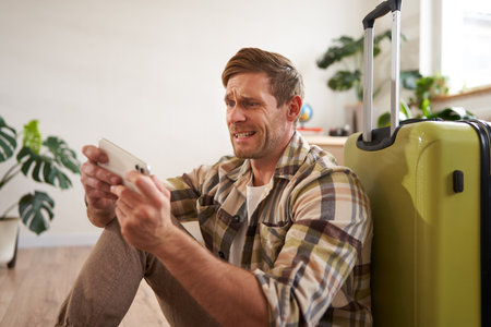 Portrait of stressed man looks at his mobile phone, playing game on smartphone, sitting near suitcase, waiting for his flight or taxiの写真素材