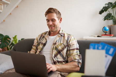 Portrait of young handsome man sitting with laptop, has passport and two plane tickets prepared, planning his journey on computer, booking hotels and restaurants online for the tripの写真素材