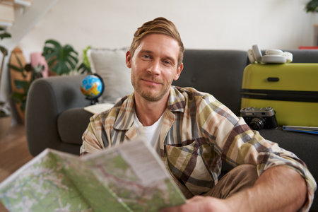 Close up portrait of smiling handsome man, boyfriend plans a holiday, holds travel map, sits near packed suitcaseの写真素材