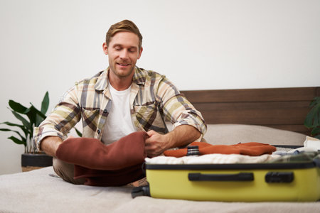 Portrait of handsome young man, tourist unpacking clothes in hotel room, staying in hostel on vacation, travelling, sitting with luggage on bedの写真素材