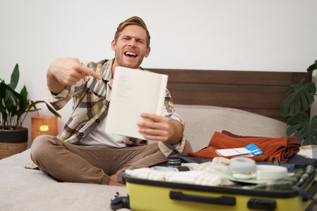Travelling and tourism. Young man sits on bed, shows his list of items, demonstrates notebook, packs clothes, prepares to go on holiday, goes on vacation, assembles luggageの写真素材