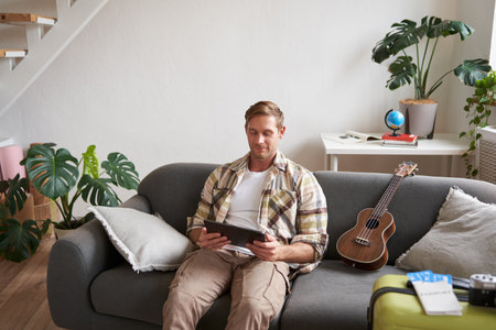 Portrait of young man in living room, sitting with tablet in hands, has suitcase packed and two plane tickets, preparing to go abroad, going on holidayの写真素材