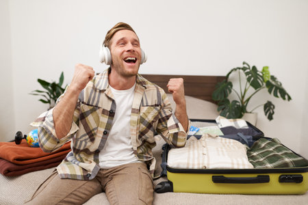 Excited young man in headphones, gets ready for vacation, goes on holiday, packs things in suitcase, listens to music in earphones, sits near his luggageの写真素材