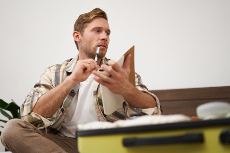 Portrait of young man, tourist looking at his notes, check-list of items to bring on vacation, packing clothes in suitcase, going on holidayの写真素材