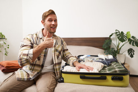 Attractive young man smiling, showing thumbs up, showing his clothes inside suitcase, getting ready for a holiday, travelling with travel agency tourの写真素材