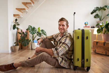 Portrait of smiling handsome man, sitting with suitcase on floor, holding passport and flight tickets, going on tour vacationの写真素材