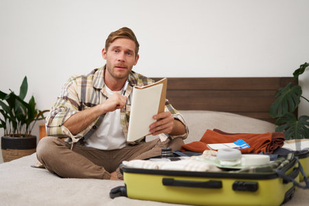 Portrait of young man, looking happy, going on holiday trip, travelling, holding notebooks with list of items, packing suitcase while sitting on bedの写真素材