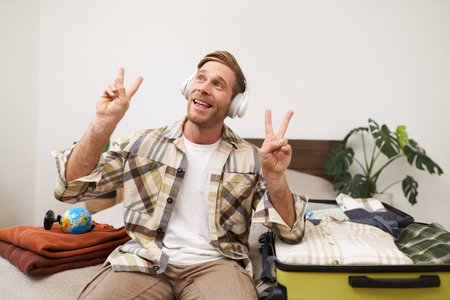 Portrait of handsome, happy young man goes on holiday, shows peace sign, listens to music in headphones, packs luggage, puts clothes in suitcase, dreams about vacationの写真素材