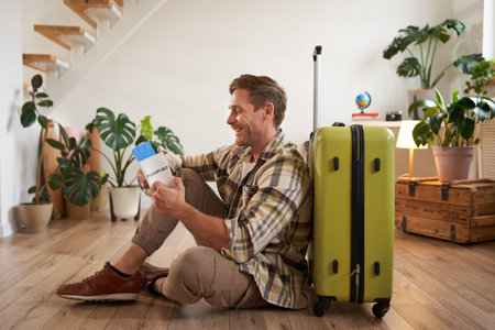 Portrait of handsome young man, tourist sitting with his suitcase, holding passport and flight tickets, going on holidayの写真素材
