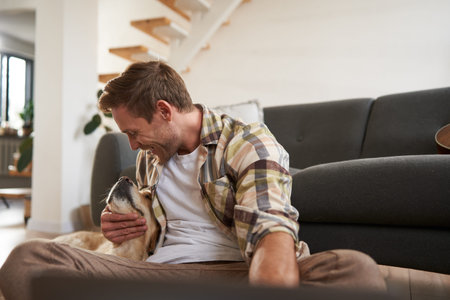 Image of happy, smiling young man playing with his dog, sitting in living room on sofa, working with laptopの写真素材