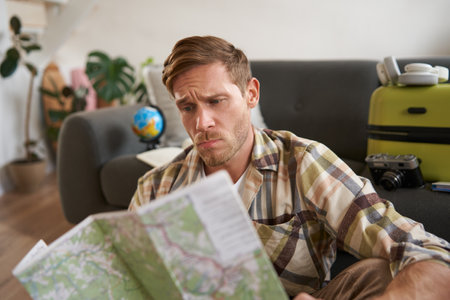 Portrait of man with concerned, puzzled face looking at map, frowning while studying the route, sitting with suitcase, going on vacationの写真素材