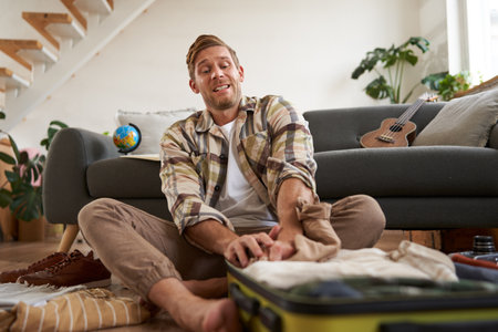 Portrait of man trying to fit clothes in full suitcase, packing luggage for holiday or business tripの写真素材
