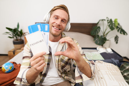 Close up portrait of handsome, happy tourist, young man shows heart sign, passport and plane tickets, sits on bed with suitcase, goes on holiday, books vacation with travel agencyの写真素材
