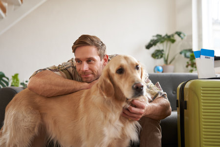 Portrait of young man hugging his dog, going on vacation to pet-friendly hotel, packed suitcase, sits in living roomの写真素材