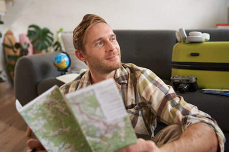 Smiling handsome man with map in hands, packed suitcase, goes on holiday, prepared to go on vacationの写真素材