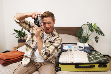 Portrait of handsome happy man, holding his analogue camera, sitting near opened suitcase, packing things for holiday, travelling abroad, getting ready for vacationの写真素材