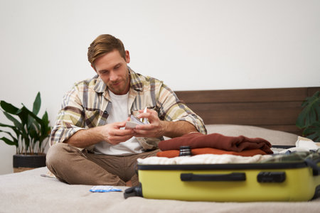 Portrait of young man with suitcase, holding his plane tickets, preparing to go on holiday, packing things for vacation, tourist puts clothes in bagの写真素材