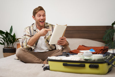 Portrait of charismatic, handsome man, tourist looking at notebook, getting ready for journey, packing suitcase, checking list of items on journalの写真素材