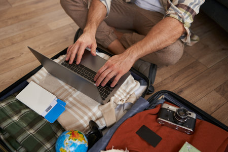 Close up shot of man, male hands using laptop on top of opened suitcase, packing for a holiday, going on vacation, prepare camera, two flight tickets for a trip abroadの写真素材