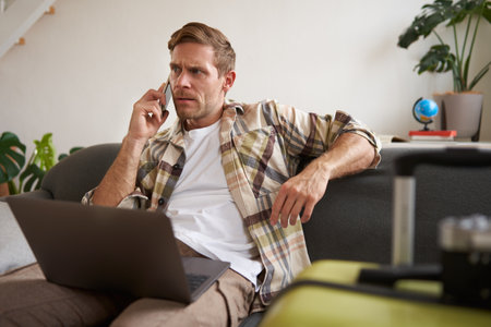 Portrait of young man with credit card, sitting and using laptop, making a phone call, booking hotel or tickets, confirming his purchase over telephone, has suitcase near himの写真素材