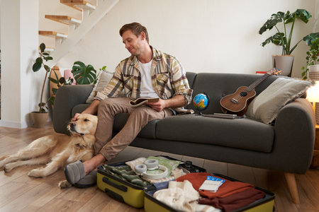 Smiling handsome guy and his dog, sitting in living room, packing suitcase for summer holiday, planning a vacation, holding notebookの写真素材