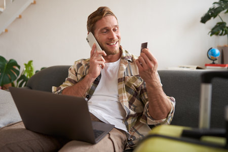 Portrait of smiling guy, tourist booking hotel, confirming purchase over the phone, holding credit card and laptopの写真素材