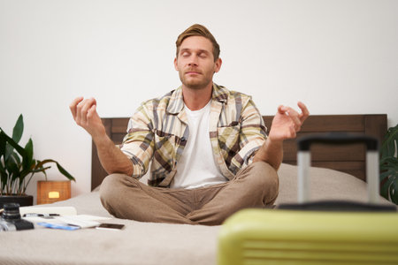 Close up portrait of meditating tourist, man sits with suitcase in bedroom, practice yoga, relaxing, finding nirvanaの写真素材