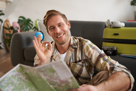 Smiling handsome tourist, man with road travel map, sits on floor near suitcase, shows okay, ok sign, recommending a tourの写真素材
