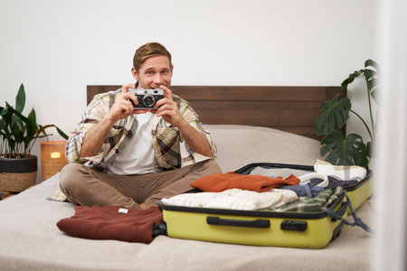 Portrait of tourist, man sits with suitcase on bed, holds camera, packs suitcase with clothes, goes on holiday, prepares for summer vacation, tourist travels abroadの写真素材