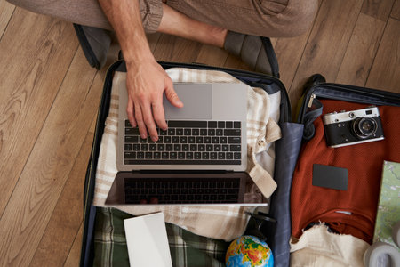 Top view of opened suitcase with clothes, male hands using laptop, booking hotel for vacation, check-in on flight for holiday, packed camera to shoot picturesの写真素材