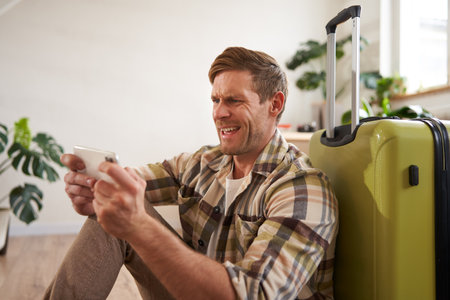 Man with shocked face, sitting on floor with suitcase, looking worried and tensed on mobile phone screenの写真素材