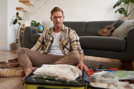 Travelling and tourism concept. Happy young man, sits on floor with luggage opened, packing suitcase for holiday, going on vacation, putting clothes inside a bagの写真素材