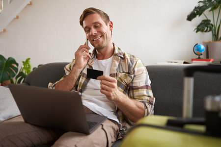 Portrait of smiling guy, tourist booking hotel, confirming purchase over the phone, holding credit card and laptopの写真素材