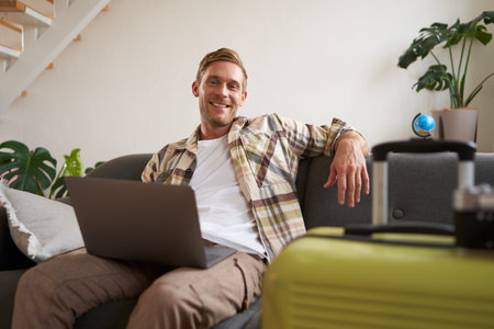 Portrait of happy young man, tourist booking his holiday with travel agency, looking at laptop, packed suitcase to go on vacationの写真素材