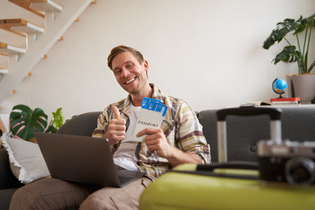 Portrait of happy handsome guy, shows his flight tickets, booked a trip for summer holiday, sits with laptop in living room, smiles at cameraの写真素材