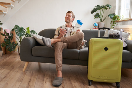 Handsome young man sits on sofa with suitcase, holds smartphone, books hostel or holiday vacation, chats on mobile phoneの写真素材