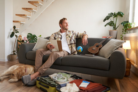 Portrait of young man sitting on sofa with notebook, has dog lying next to him, packing suitcase for summer vacationの写真素材
