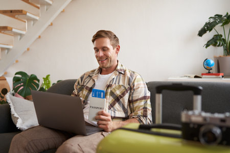 Portrait of handsome young man, has two tickets and passport, goes online on laptop, booking hotel and planning his holiday, sitting in living roomの写真素材