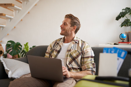 Portrait of handsome young man, has two tickets and passport, goes online on laptop, booking hotel and planning his holiday, sitting in living roomの写真素材