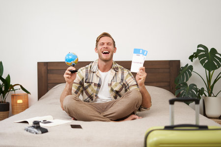 Portrait of cheerful, happy tourist, man sitting with plane tickets and a globe, packed suitcase for travelling around the world, going on vacation, excited about his holidayの写真素材