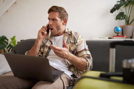 Portrait of angry man arguing over the phone, has laptop opened on his laptop, sits near suitcase, complaining at travel agencyの写真素材