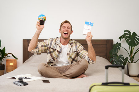 Portrait of cheerful, happy tourist, man sitting with plane tickets and a globe, packed suitcase for travelling around the world, going on vacation, excited about his holidayの写真素材