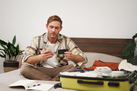 Portrait of happy man in suitcase, transferring money, buying tickets online, sitting on bed with credit card and smartphone app, paying onlineの写真素材