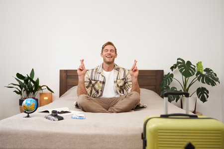Young man with globe and camera, sits on bed and meditating, has packed suitcase standing near him, going on vacation, praying for good weather on holiday, making wish with fingers crossedの写真素材