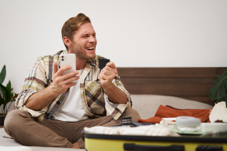 Cheerful male tourist, man packing suitcase, paying for tickets with credit card and smartphone app, celebrating, excited about going on holidayの写真素材