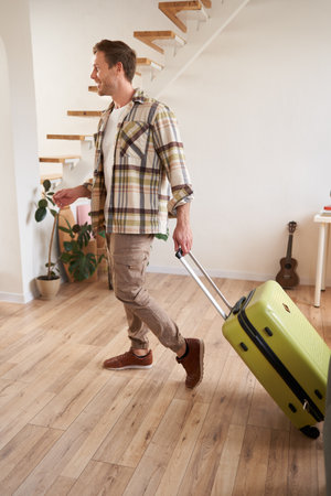 Full length photo of young man with suitcase, going on holiday trip, walking in flat, checking out from rented apartmentの写真素材