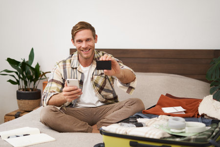 Portrait of happy man buying tickets with credit card cashback, sitting on bed and packing clothes for holiday, putting items in suitcase, holding mobile phoneの写真素材