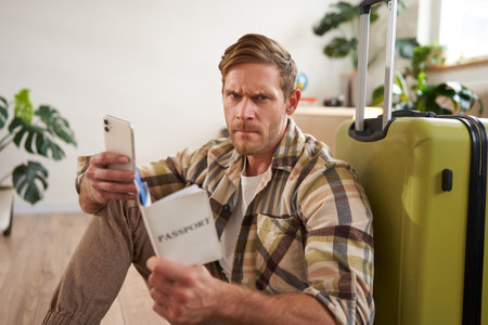 Angry man sits with suitcase, holds mobile phone and passportの写真素材