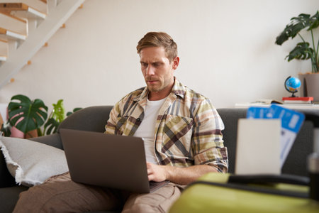 Portrait of serious and concentrated man, sits with laptop in living room, has two plane tickets, plans holiday, goes onlineの写真素材